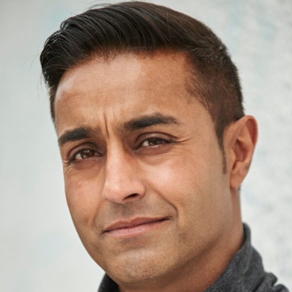 mohan-randhawa-1 A headshot of a middle-aged man with dark hair wearing a grey turtleneck against a textured background.
