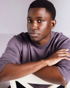 A personable Black male model with short dark hair wears a purple sweater against a studio background.