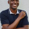 deondre-cumberbatch-13 A friendly Black male model with a warm, genuine smile, wearing a dark polo shirt against a wall.