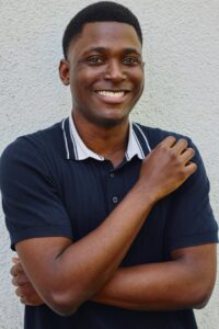 A friendly Black male model with a warm, genuine smile, wearing a dark polo shirt against a wall.