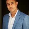 mohan-randhawa-2 A professional middle-aged male model wearing a light blue blazer against a simple dark backdrop.