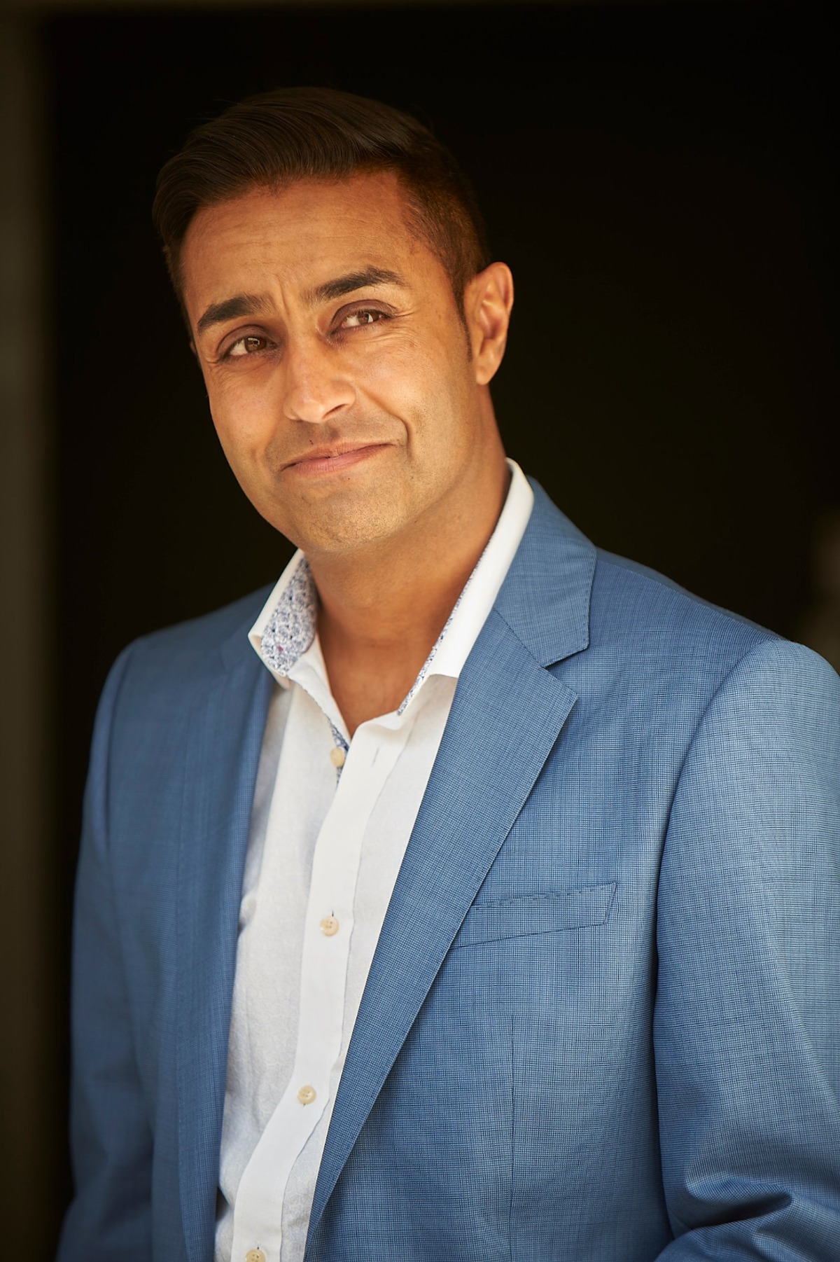 A professional middle-aged male model wearing a light blue blazer against a simple dark backdrop.