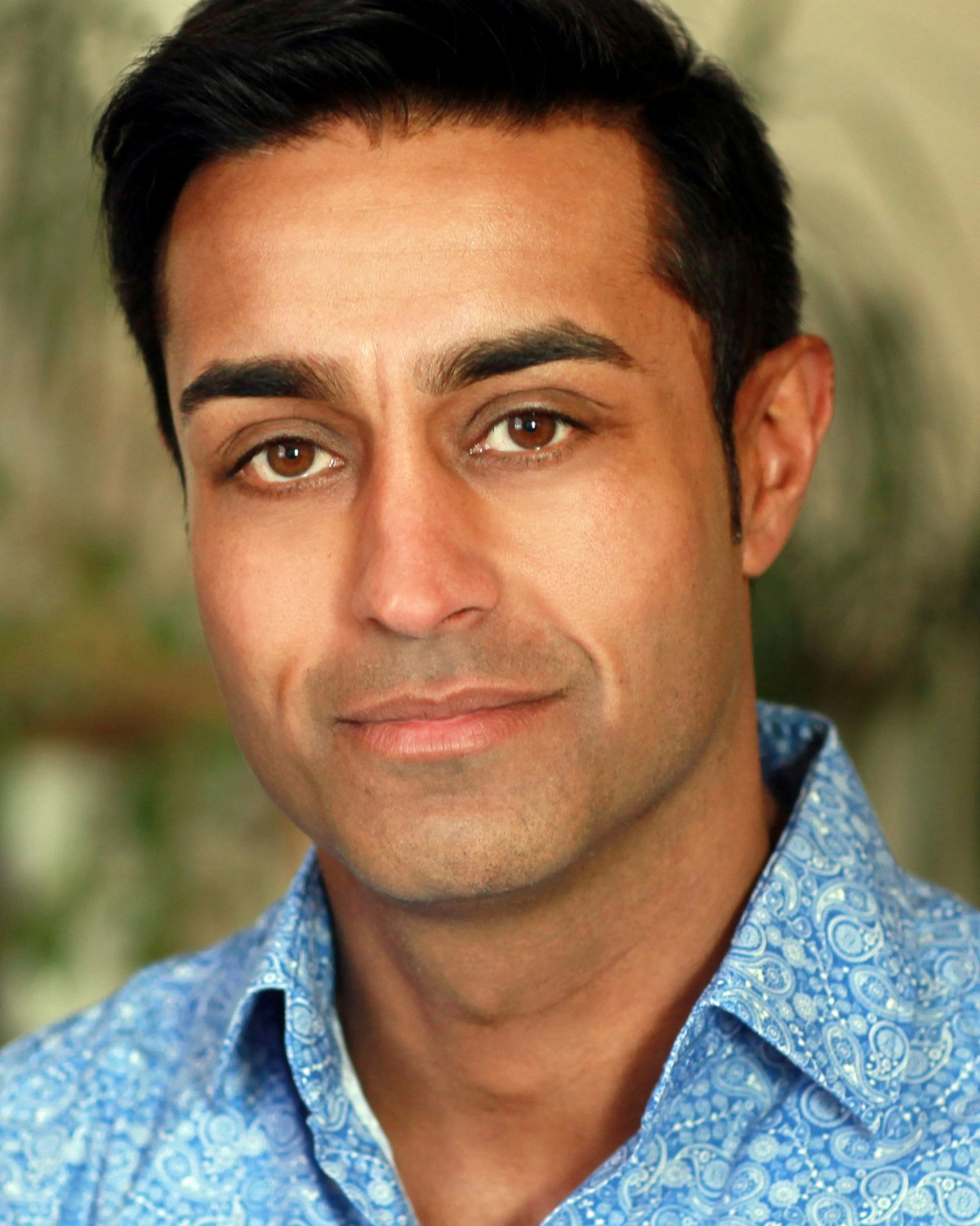 A warm portrait of a man with dark hair wearing a blue patterned shirt against blurred greenery.