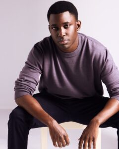 A man wearing a mauve sweater sits against a plain background, looking directly into the camera lens.