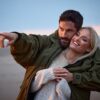 A happy couple enjoys a sunset beach walk, with the man pointing forward as they embrace together.