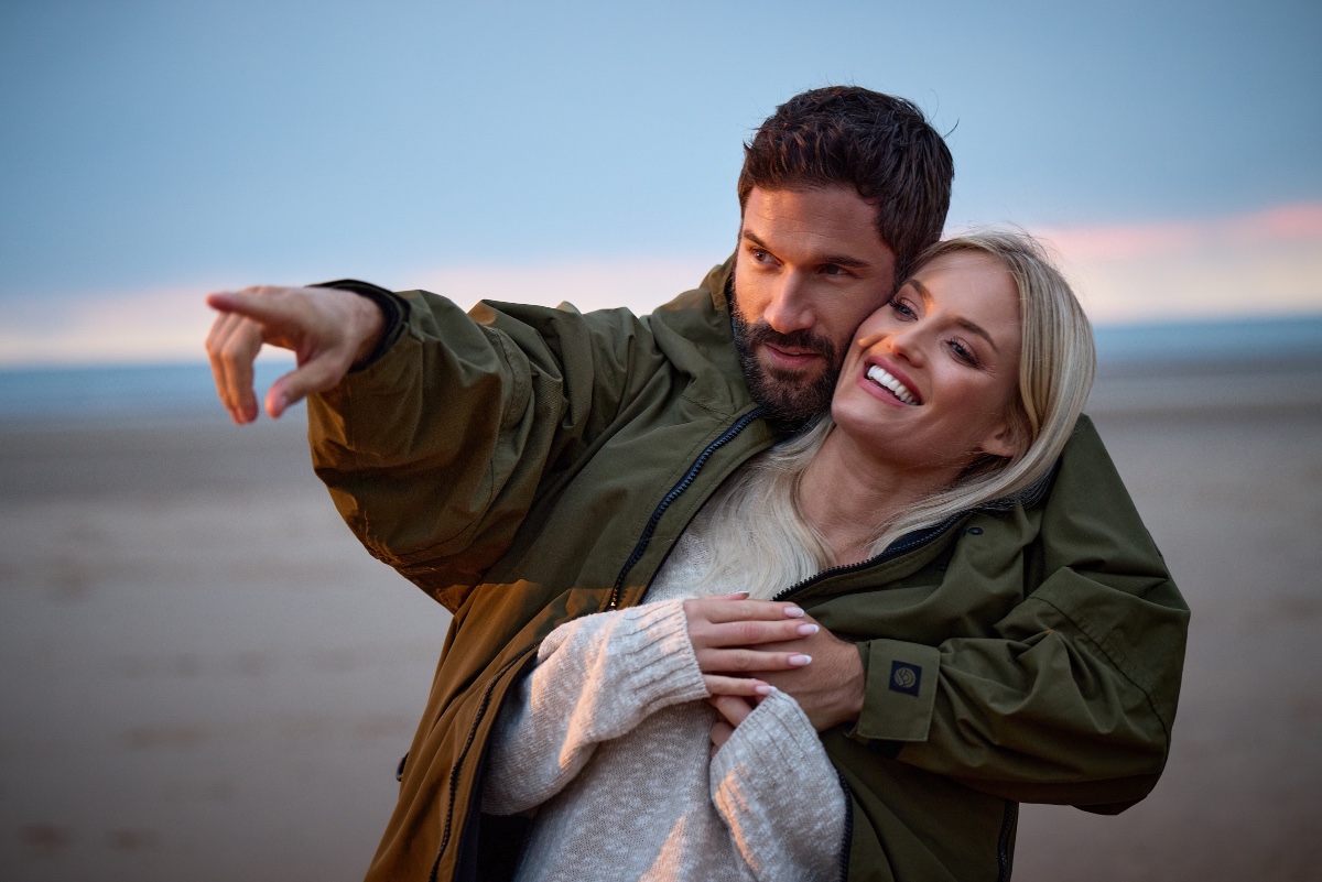 A happy couple enjoys a sunset beach walk, with the man pointing forward as they embrace together.