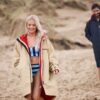 A smiling blonde woman wearing a branded dry robe on a beach with a man walking behind.