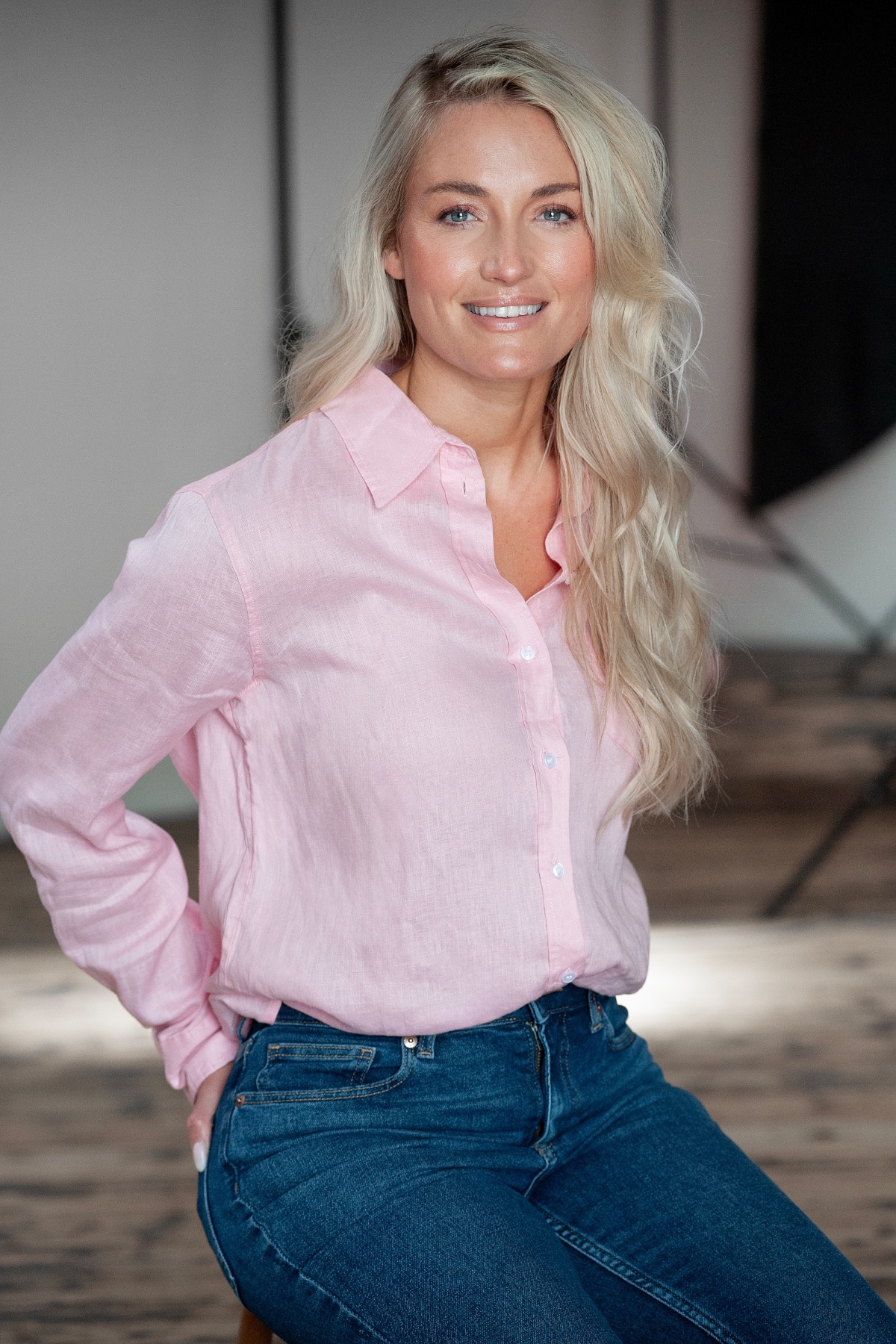 A blonde woman in a pink shirt smiling warmly, captured in a clean, brightly lit professional studio setting.