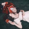 A couple in period theatrical costumes lie on the grass in a romantic, cinematic reenactment scene.