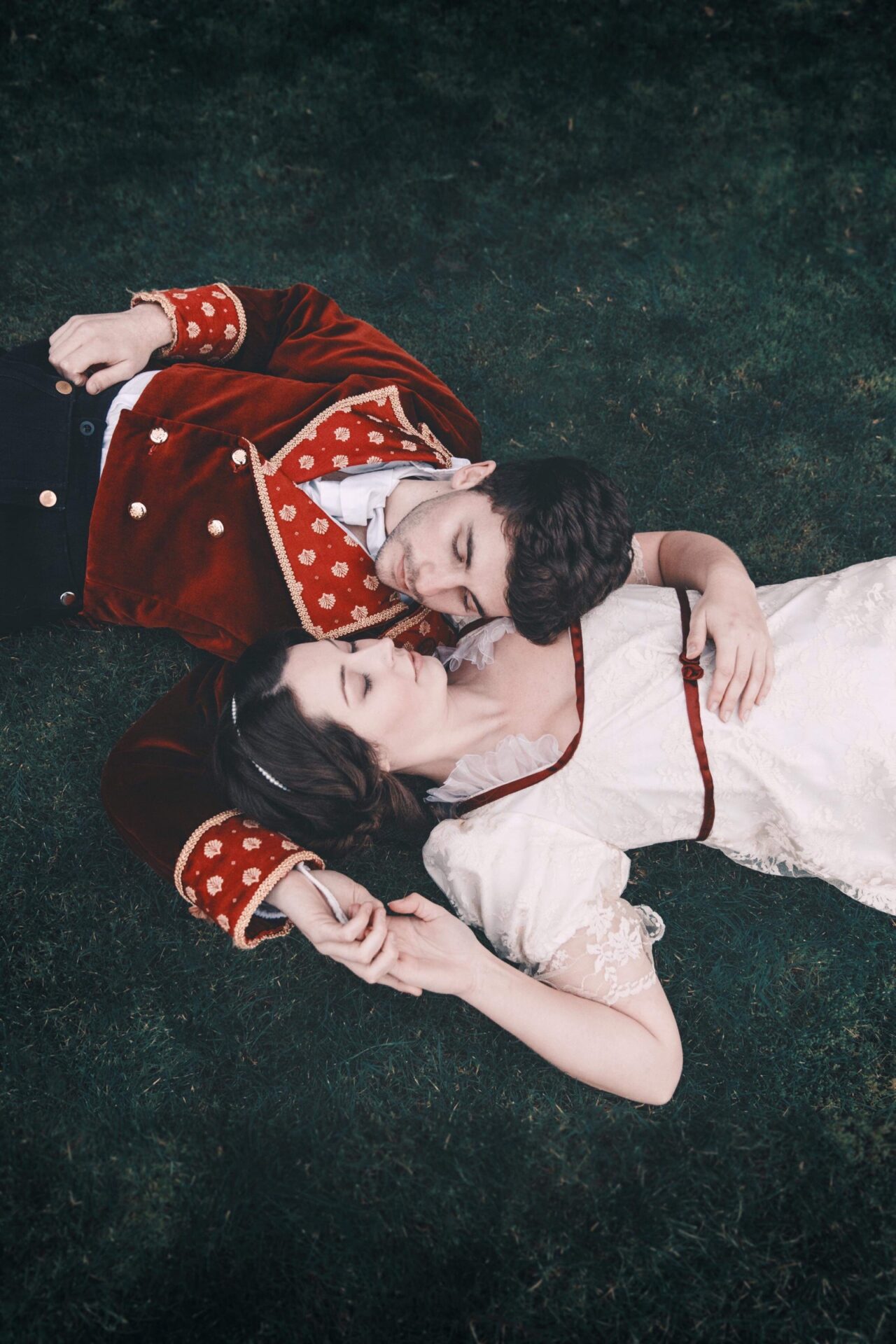 A couple in period theatrical costumes lie on the grass in a romantic, cinematic reenactment scene.
