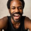 nathan-lobban-4 A charismatic Black man with a beard laughing heartily, showing bright energy against a neutral grey background.