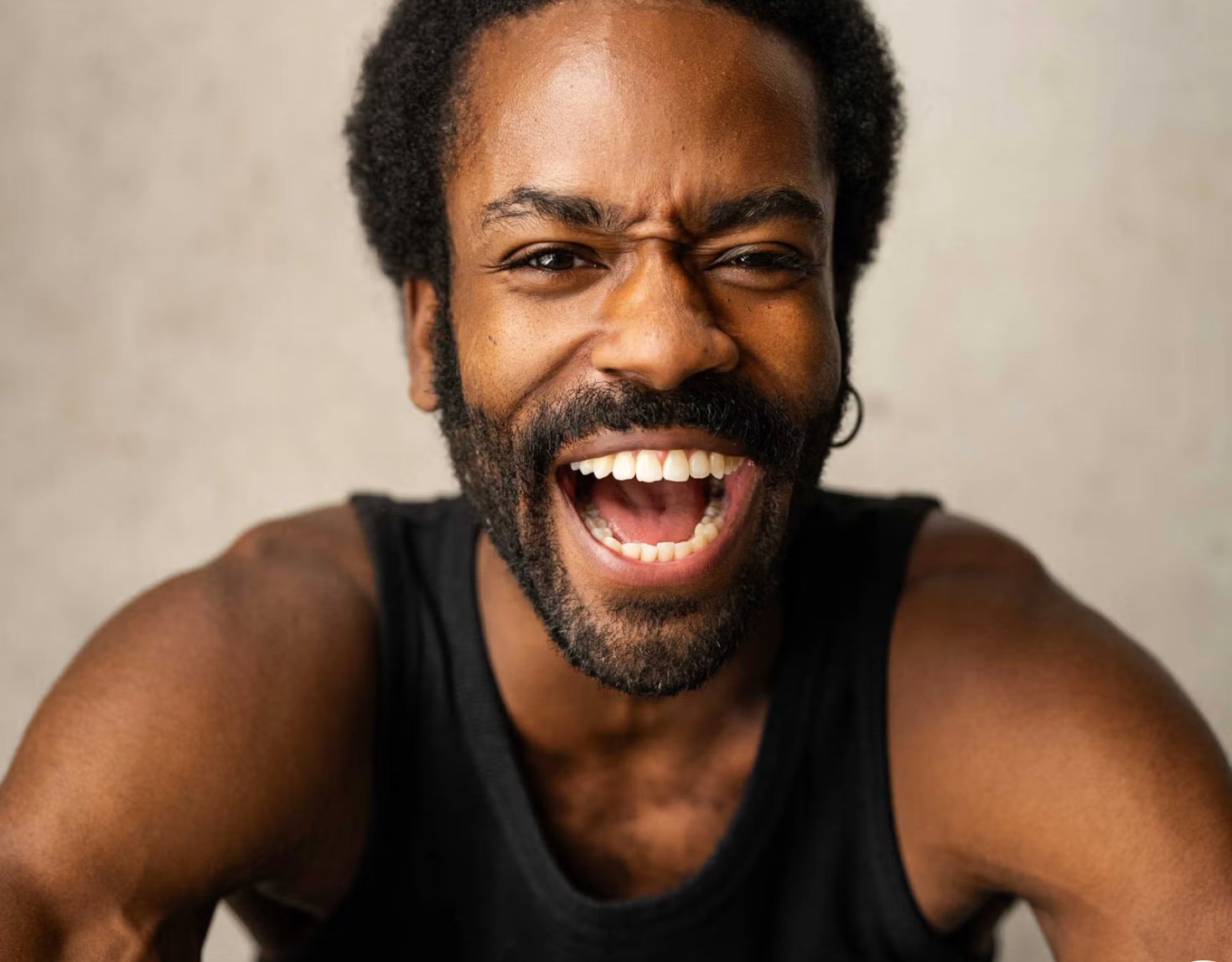 A charismatic Black man with a beard laughing heartily, showing bright energy against a neutral grey background.