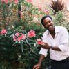 nathan-lobban-7 A smiling man in a striped shirt stands in a bright garden beside pink flowering shrubs.