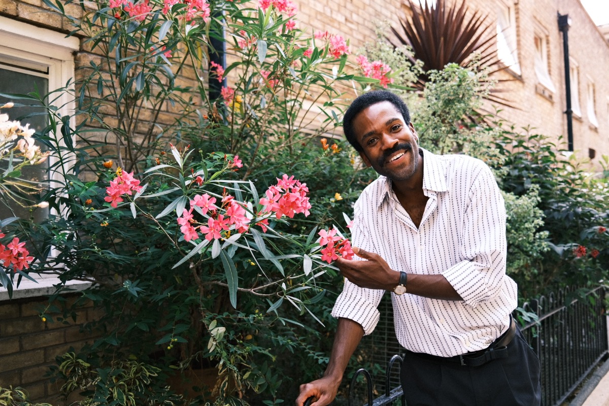 A smiling man in a striped shirt stands in a bright garden beside pink flowering shrubs.