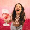A joyous woman holding a wine glass prop against a vibrant pink paper-circle background, laughing with energy.