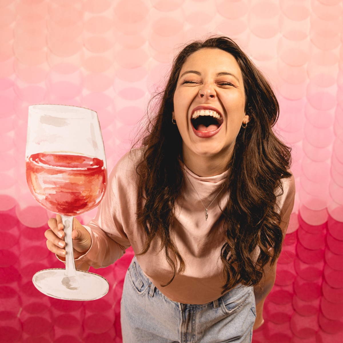 A joyous woman holding a wine glass prop against a vibrant pink paper-circle background, laughing with energy.