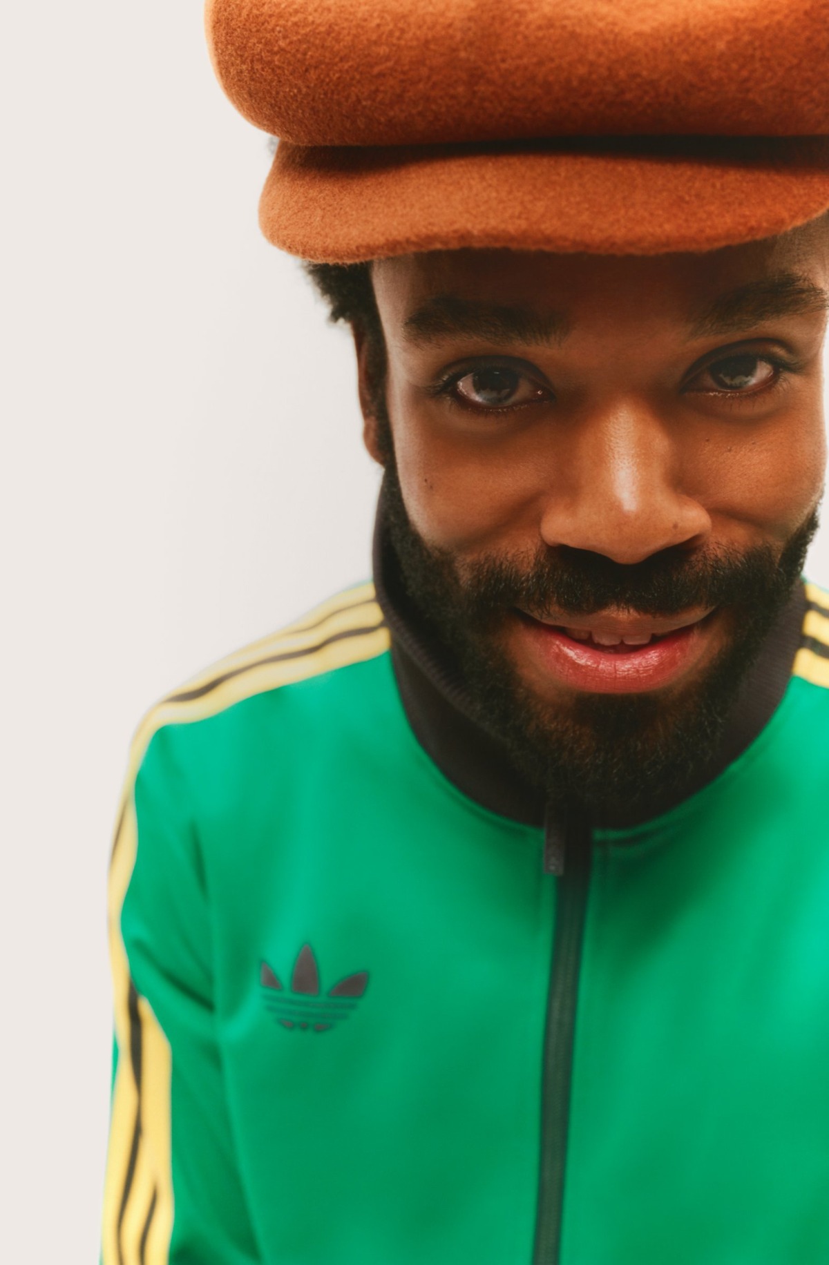 A man wearing a brown hat and green vintage tracksuit smiles warmly against a clean white background.