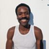 nathan-lobban-3 A smiling man with a moustache wearing a white tank top poses against a bright white wall.