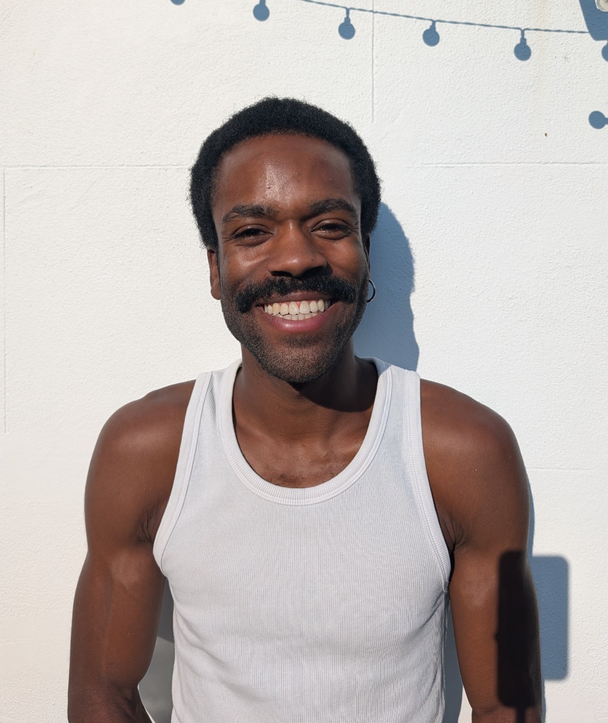 A smiling man with a moustache wearing a white tank top poses against a bright white wall.