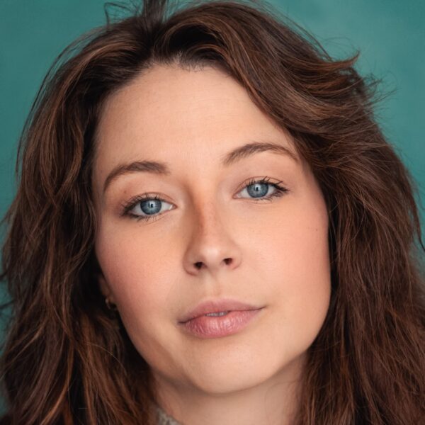 A headshot of a woman with long wavy brown hair against a textured teal studio background.