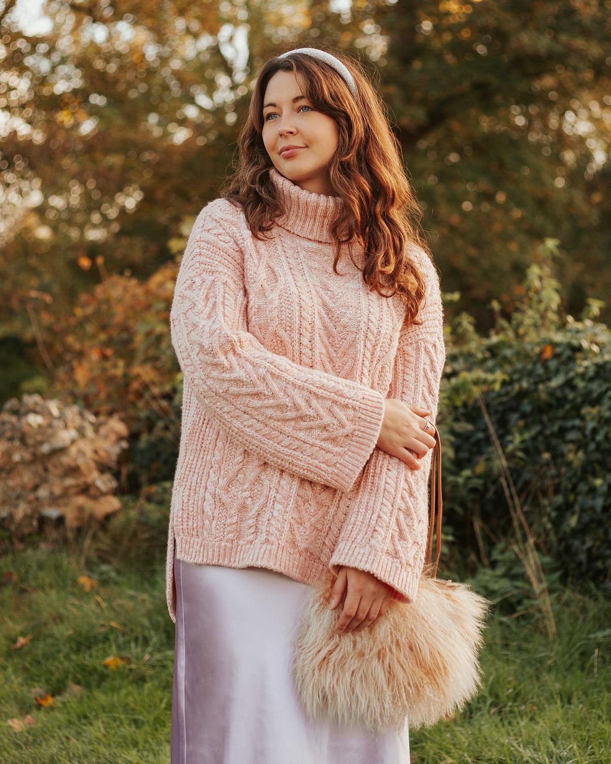 A woman wearing a cosy pink jumper and satin skirt stands outside in a golden autumn setting.