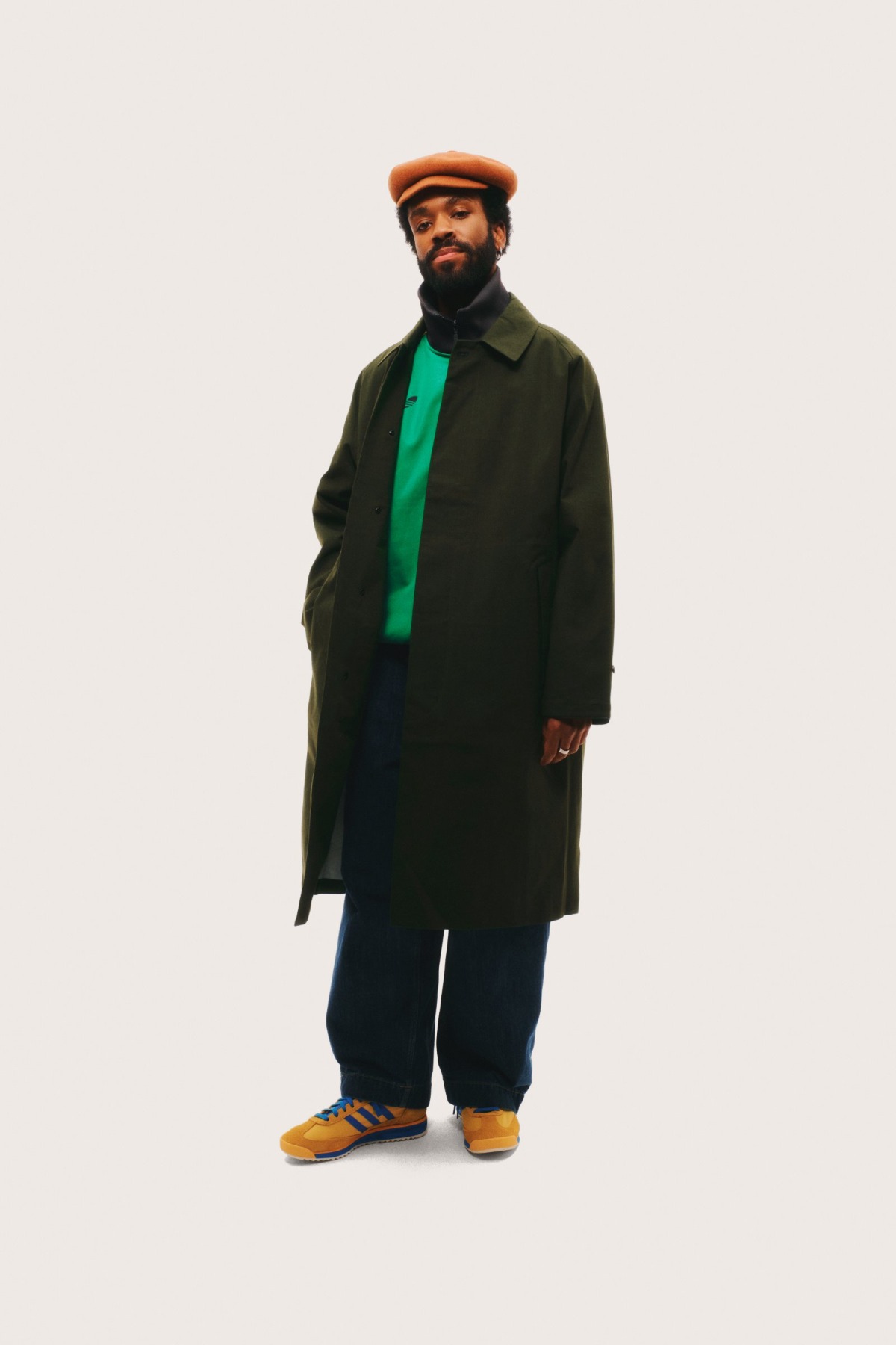 A stylish bearded man stands against a plain background, wearing a green coat, cap, and blue jeans.