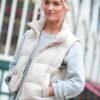 A blonde model wearing a cream puffer gilet and grey sweater, stands outdoors in a bright, casual setting.