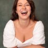 A cheerful brunette woman with a bright, genuine smile wearing a white blouse against a green background.