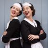 Two models dressed in vintage maid costumes laugh together while standing back to back against a door.