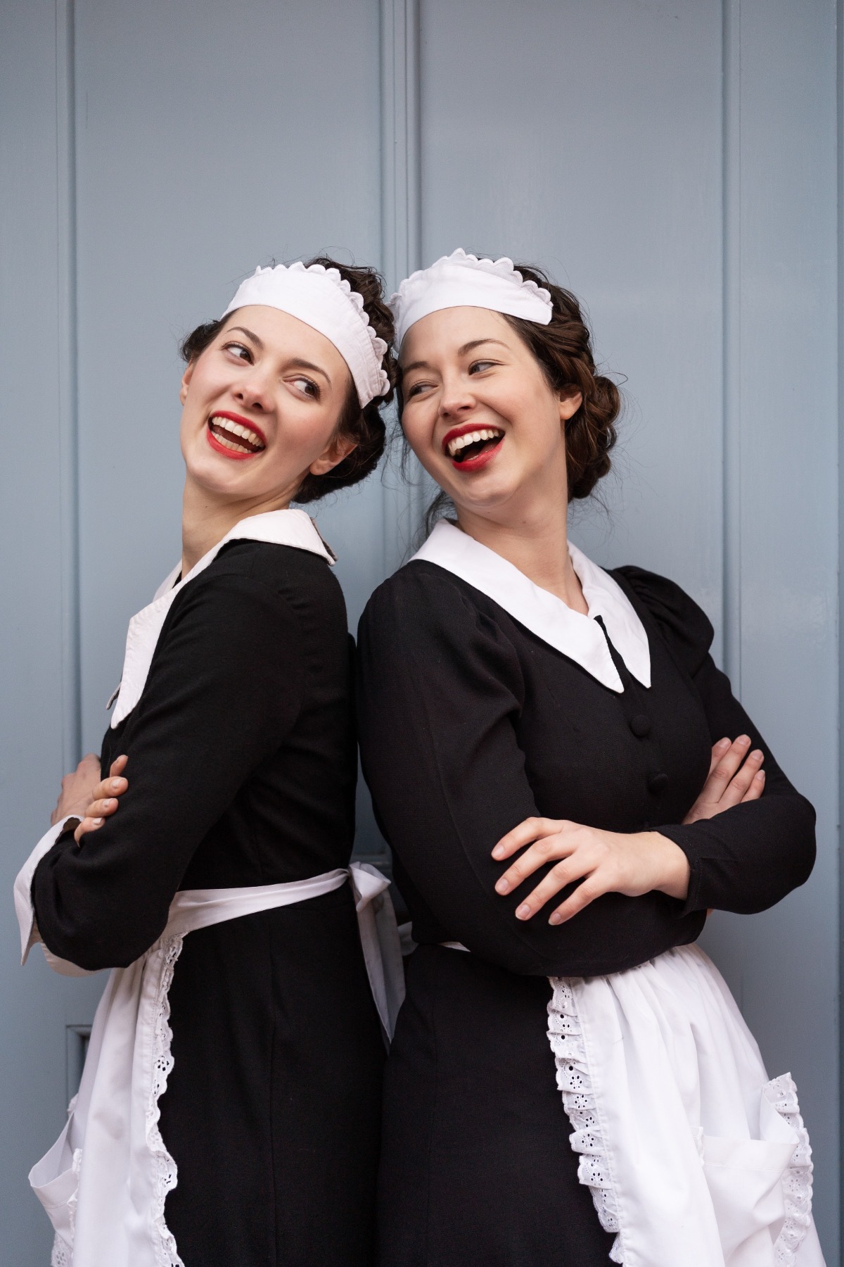 Two models dressed in vintage maid costumes laugh together while standing back to back against a door.