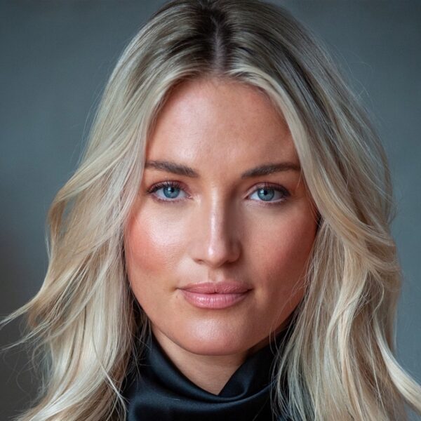 A blonde woman with blue eyes poses for a professional front-facing studio headshot against a grey backdrop.