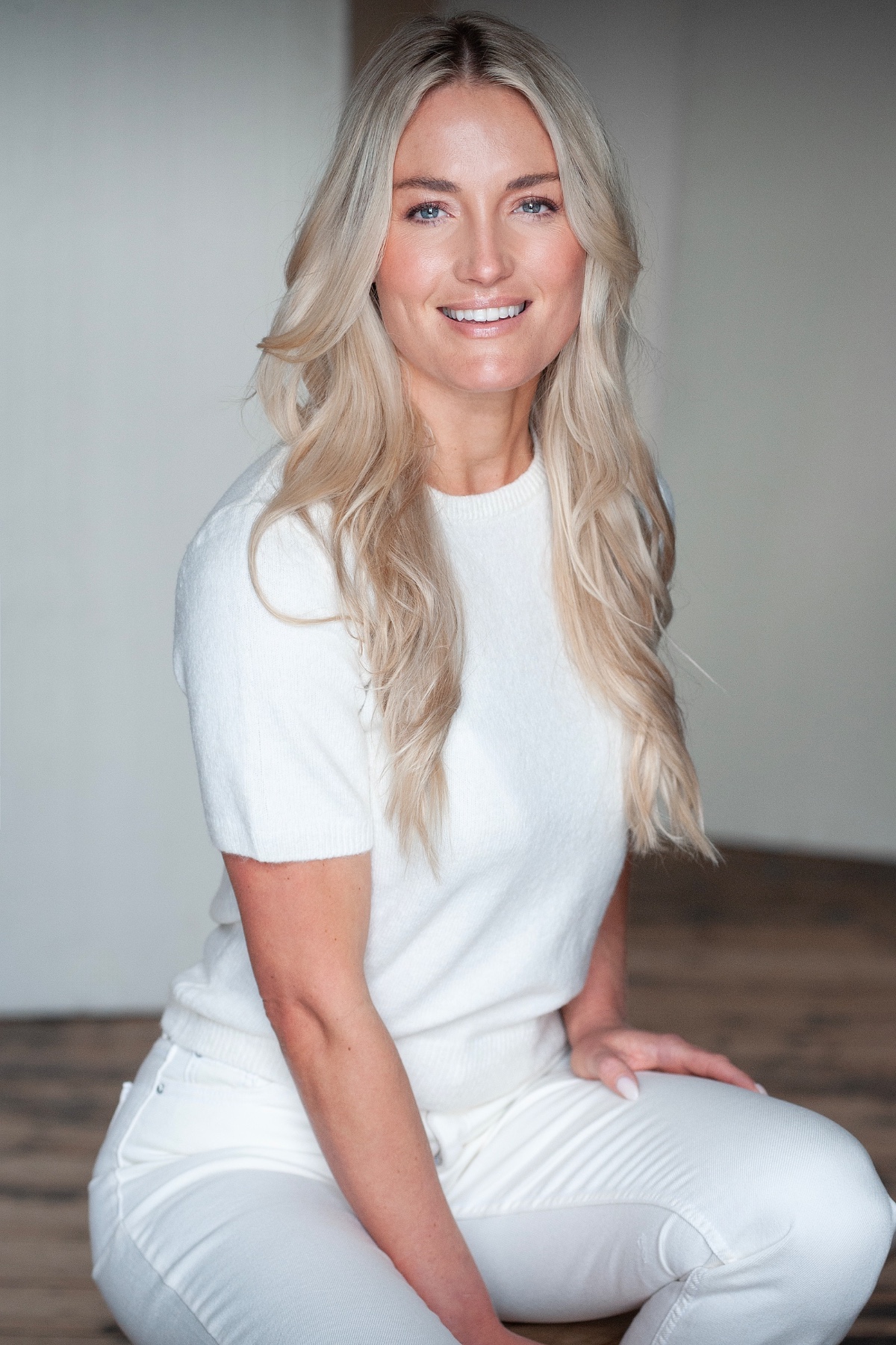 A smiling blonde woman wearing a simple white jumper, posing for a clean and professional studio headshot.