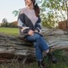 A smiling young woman in a chunky knit sweater sits on a rustic log during an autumn walk.