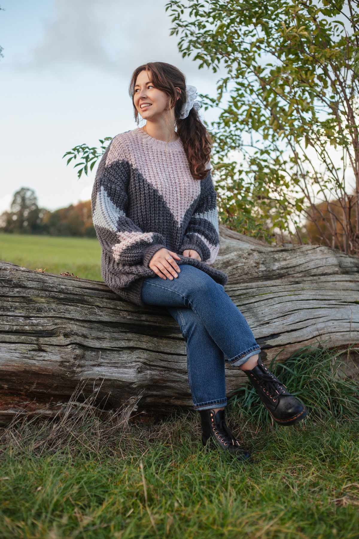 A smiling young woman in a chunky knit sweater sits on a rustic log during an autumn walk.