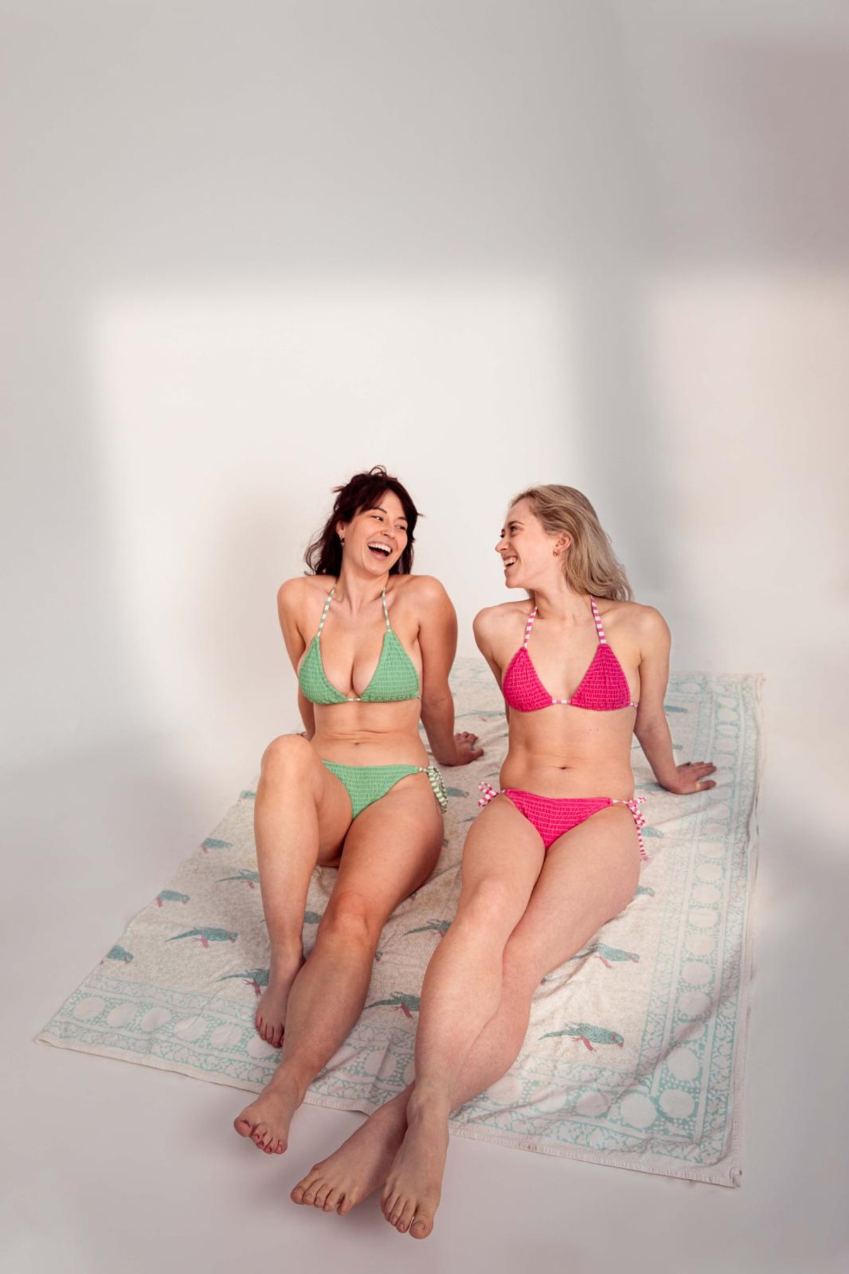 Two diverse female models wearing colourful crochet bikinis laugh together on a beach towel in studio lighting.