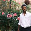 nathan-lobban-9 A stylish Black man poses naturally in a sunlit garden, wearing a patterned shirt against floral greenery.
