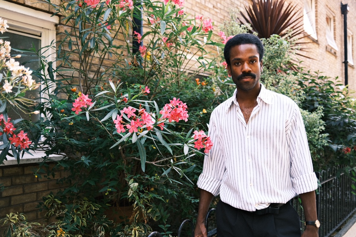 A stylish Black man poses naturally in a sunlit garden, wearing a patterned shirt against floral greenery.