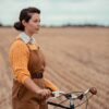 A woman in a yellow sweater and brown overalls stands with a bicycle in a rural field.