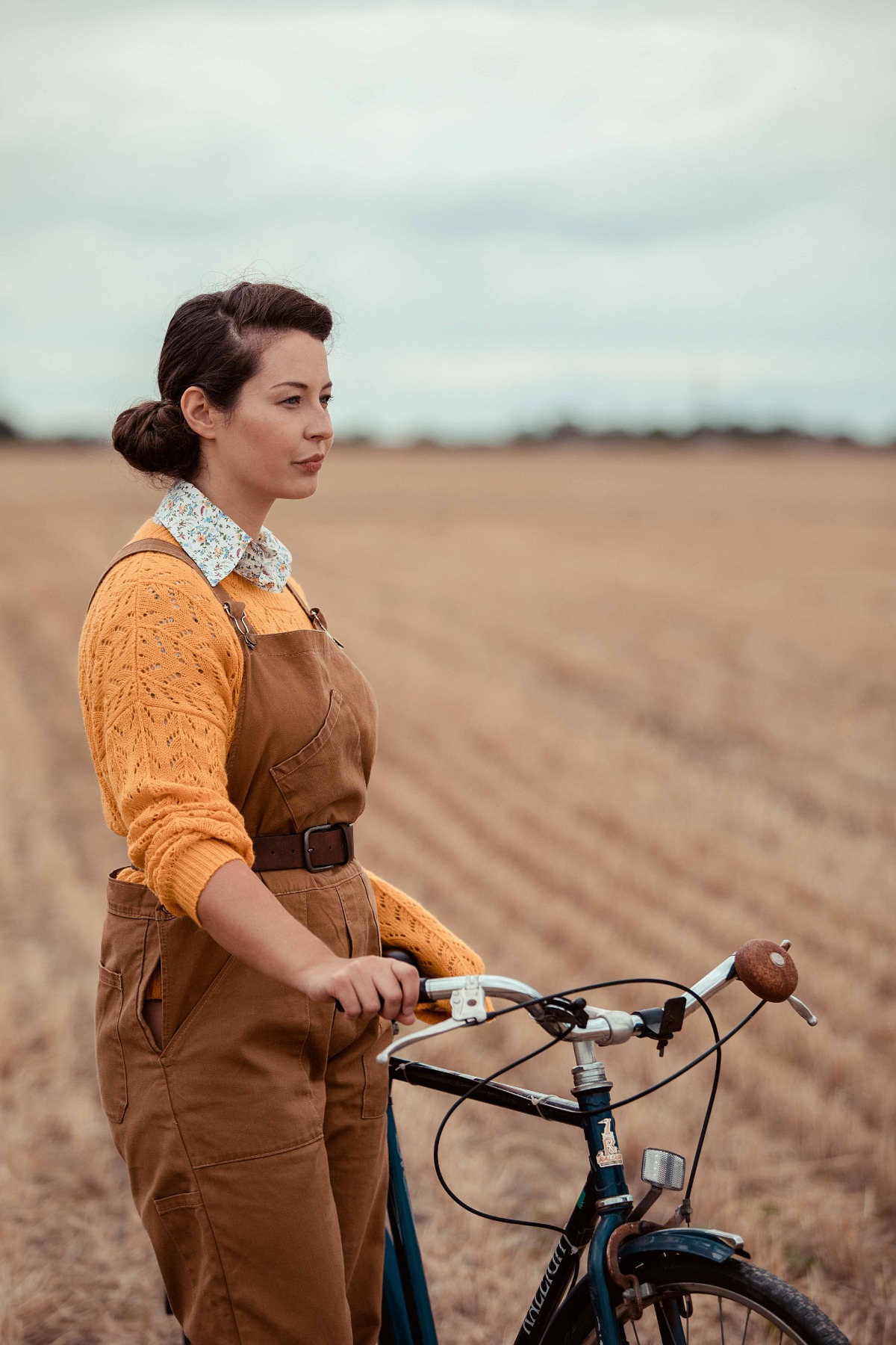A woman in a yellow sweater and brown overalls stands with a bicycle in a rural field.