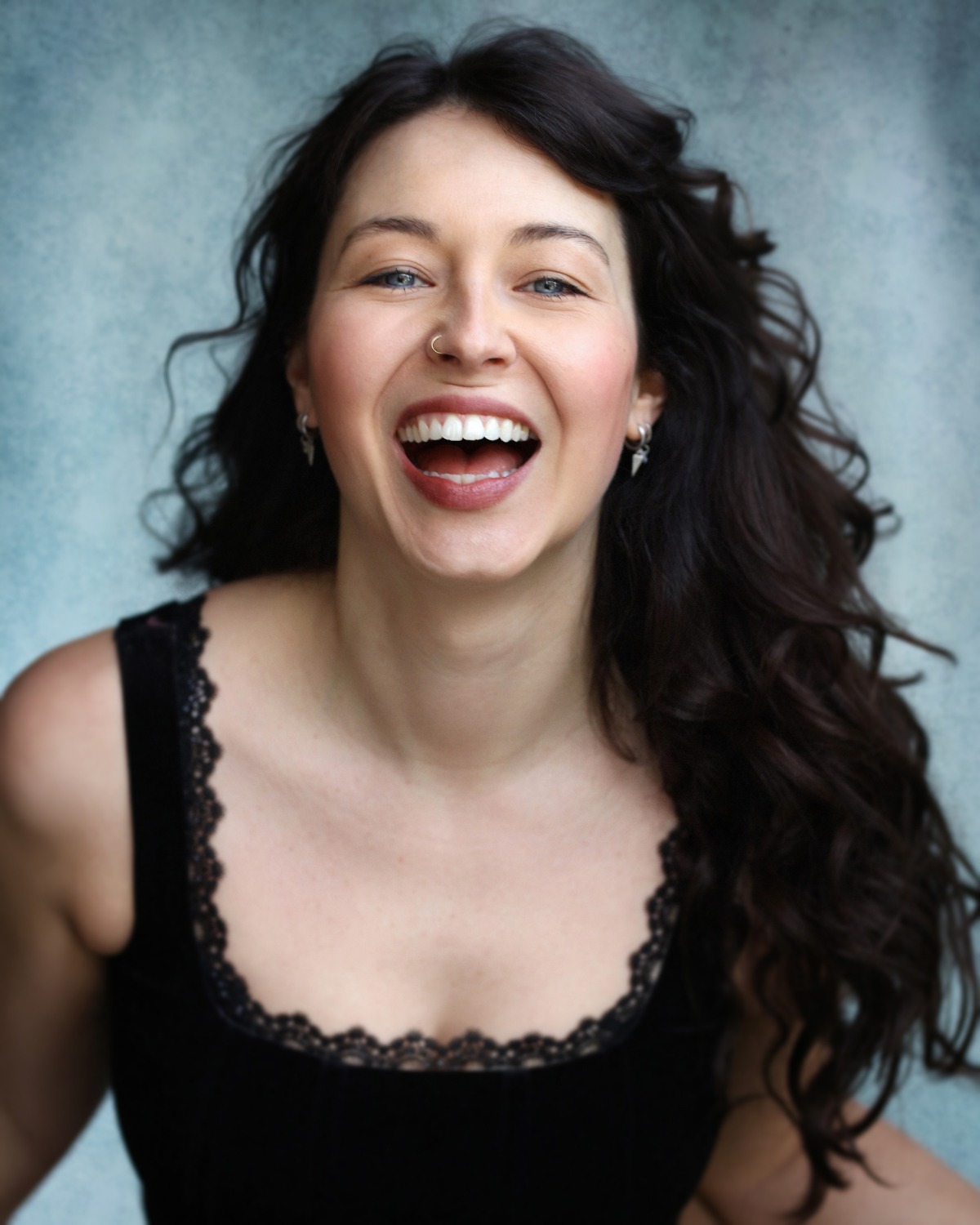 A charismatic woman with long dark wavy hair smiles broadly against a textured blue studio backdrop.