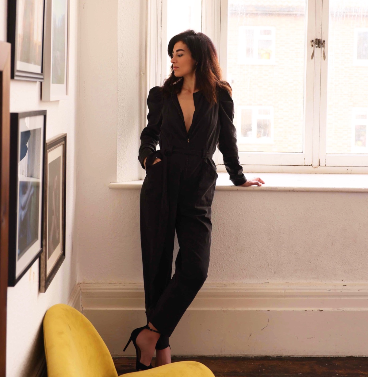 helena-antonio-8 A stylish brunette woman in a black jumpsuit leans against a window in a bright living space.