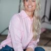 A blonde woman in a pink linen shirt smiles warmly while posing in a bright, modern studio.