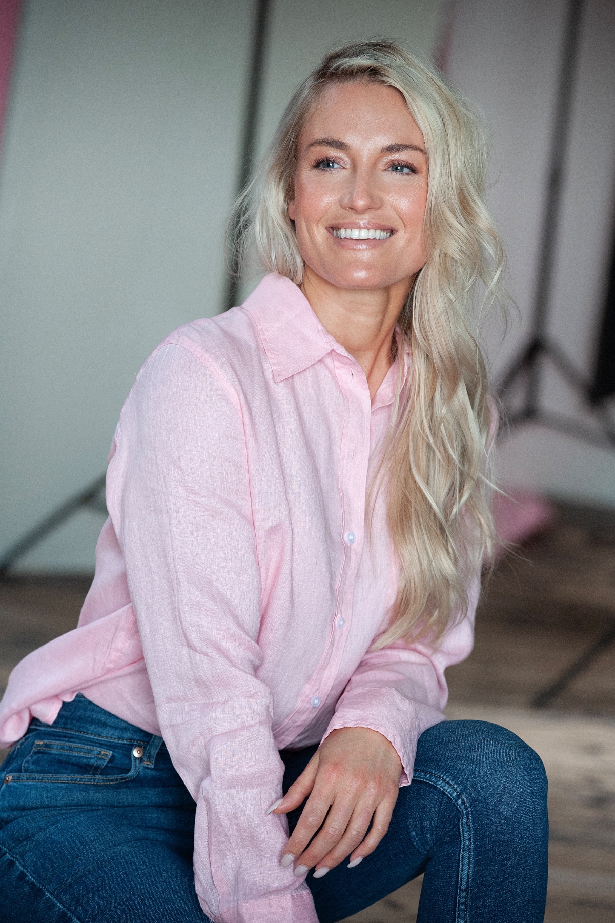 A blonde woman in a pink linen shirt smiles warmly while posing in a bright, modern studio.