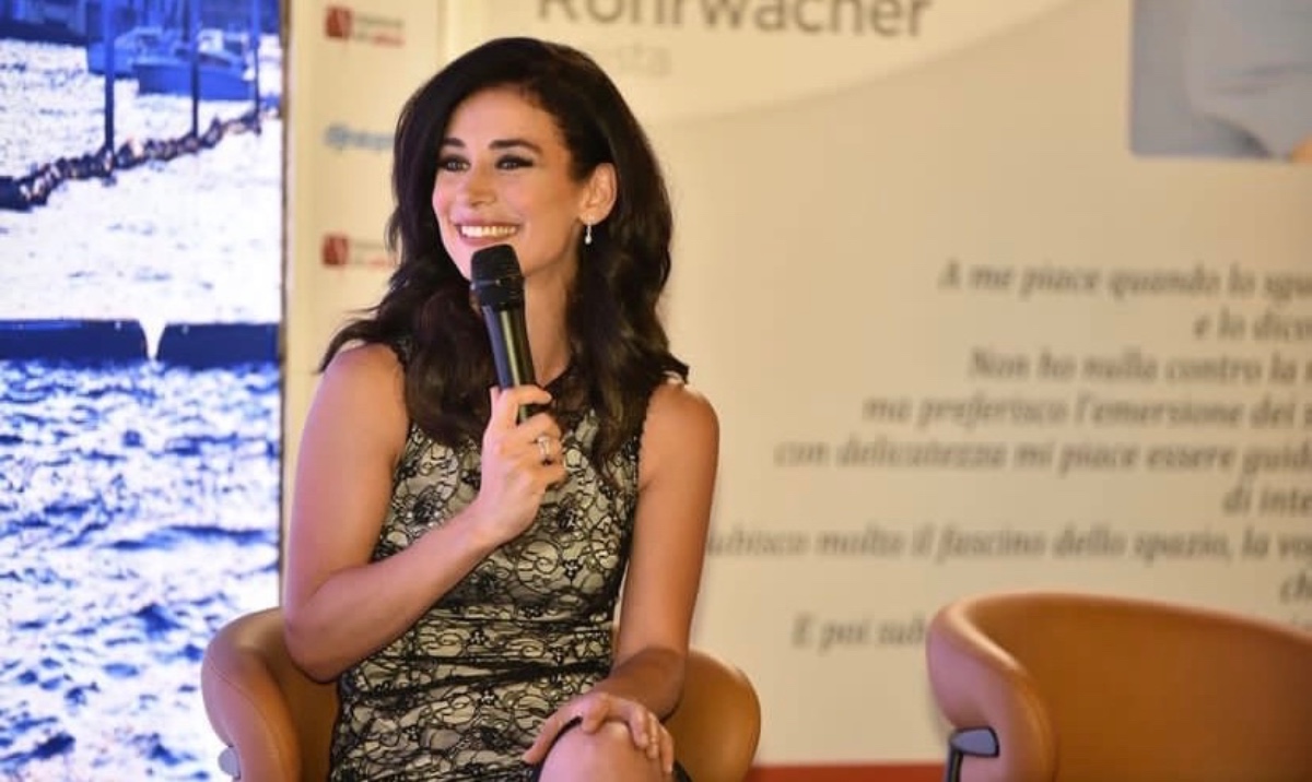 helena-antonio-9 A smiling woman in a lace dress speaks into a microphone during a professional event or presentation.
