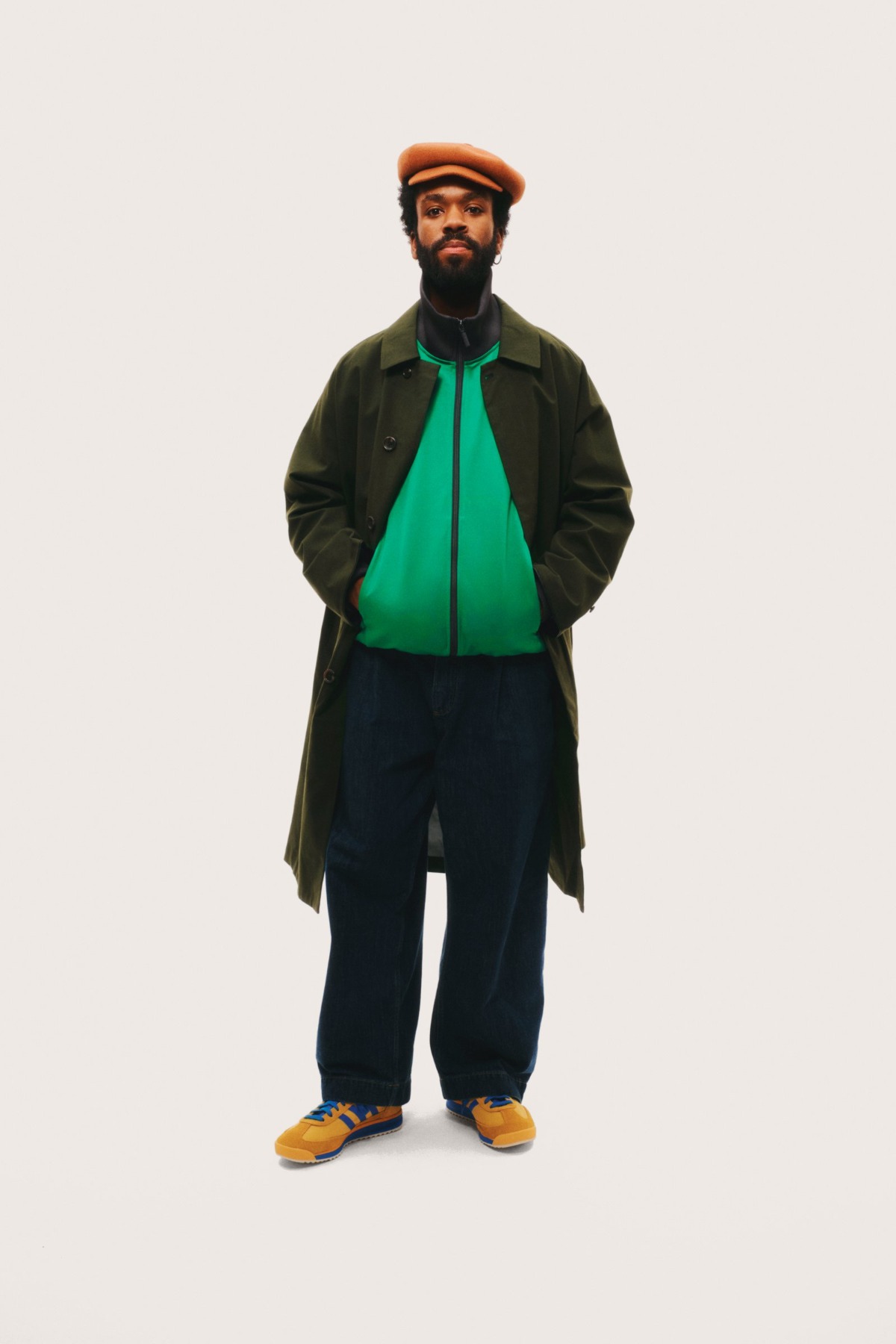 A stylish bearded man stands against a plain background wearing a green jacket, coat, and orange cap.