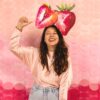 A radiant woman laughing and holding a strawberry prop in front of a bright pink textured backdrop.