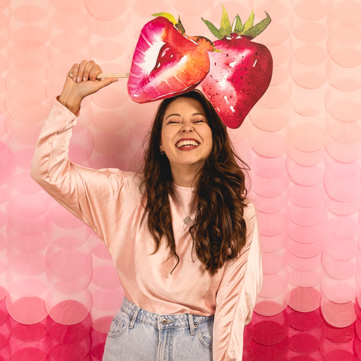 A radiant woman laughing and holding a strawberry prop in front of a bright pink textured backdrop.