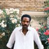 nathan-lobban-8 A stylish Black male model with a mustache poses in a garden, wearing a casual striped shirt outdoors.