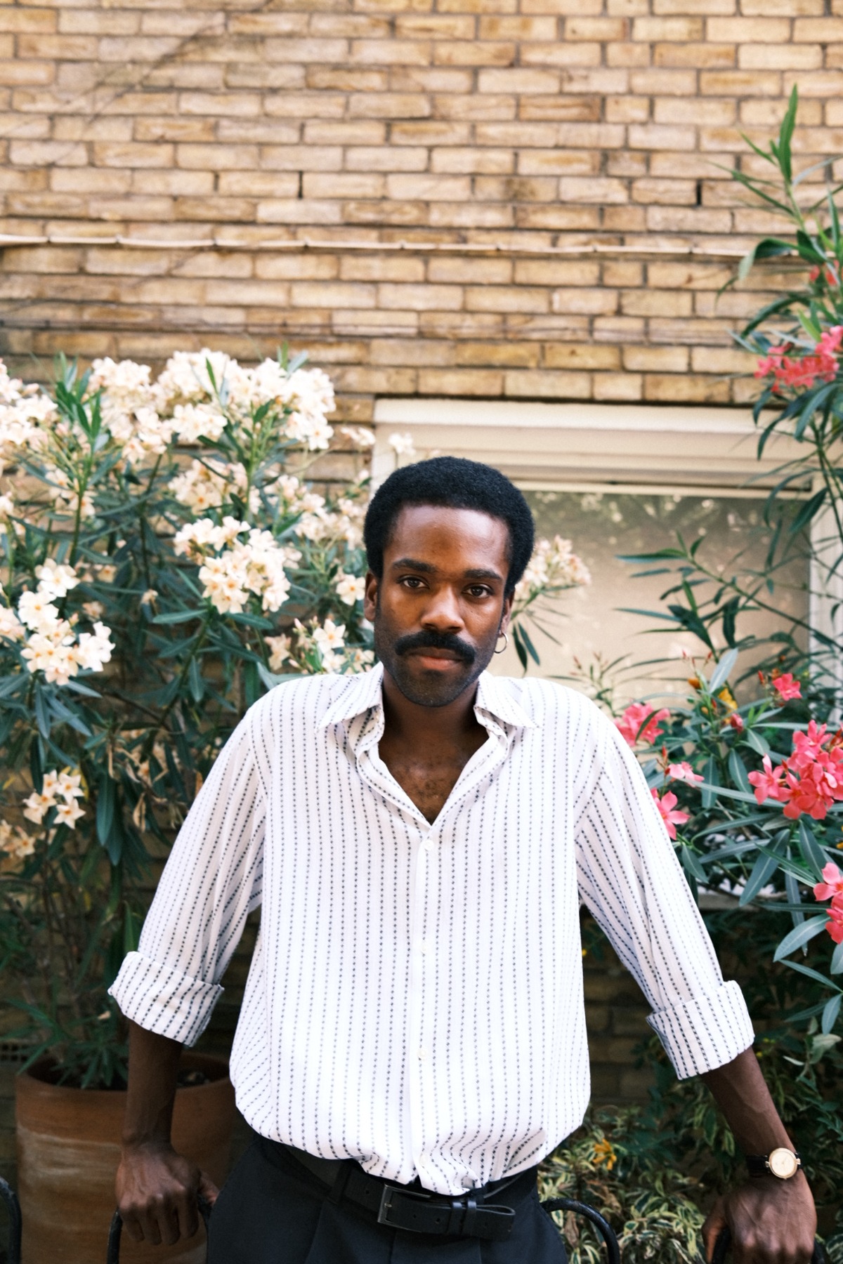 A stylish Black male model with a mustache poses in a garden, wearing a casual striped shirt outdoors.