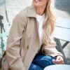 A smiling blonde woman wearing a trench coat sits outdoors at a cafe table, looking away thoughtfully.